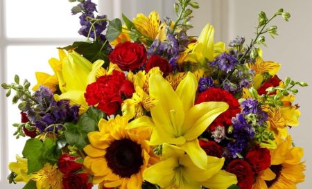 Sunlit yellow Asiatic Lilies, sunflowers, red carnations, red spray roses, yellow Peruvian Lilies, purple larkspur, and ivy vines are professionally designed in a clear glass bowl vase to express your floral sentiment.